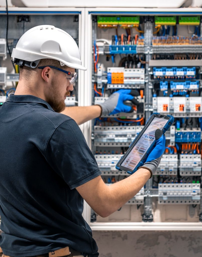 Electrical technician working in a switchboard with fuses, uses a tablet.